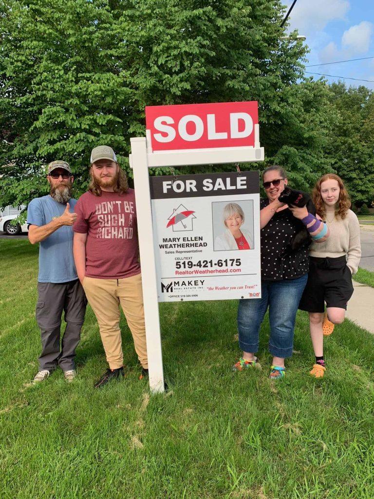 Alt text: Family celebrating home sale, standing next to realtor under a large "SOLD" sign.